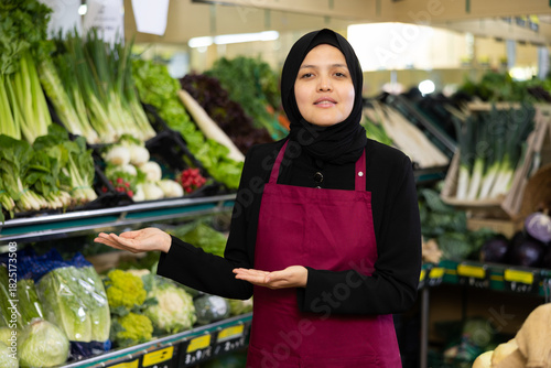 Female seller in a hijab and an apron offers to buy vegetables and fruits in the grocery department of a supermarket
