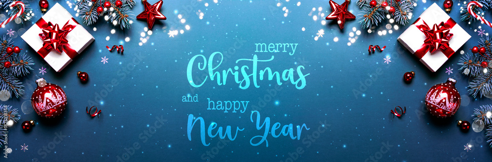 Fototapeta premium Merry Christmas and Happy New Year text on blue background with gift boxes, ribbons, red decoration, fir branches, bokeh, sparkles. Xmas and winter holidays. Christmas banner, top view