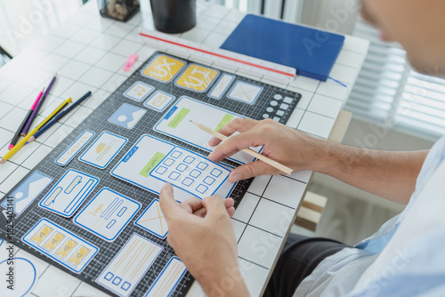 A designer is developing UX UI layouts, reviewing mobile app prototypes, and arranging interface elements on paper and board. He is focused on user experience and digital creativity.