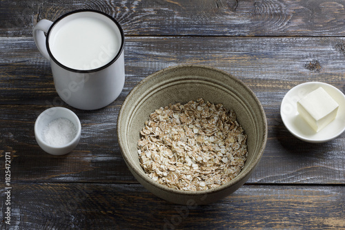 Raw four grain flakes in a ceramic bowl with milk, butter and salt arranged on a rustic wooden table, showing natural ingredients for healthy porridge