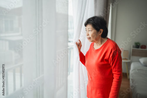 Thoughtful elderly woman in a bright room gazing out the window, reflecting on life and memories, wearing a cozy red sweater, with soft natural light filtering through drapes