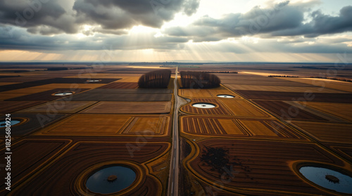 Stunning aerial landscape of agricultural fields with circular irrigation systems and a straight road, bathed in dramatic sun rays piercing through a cloudy sky