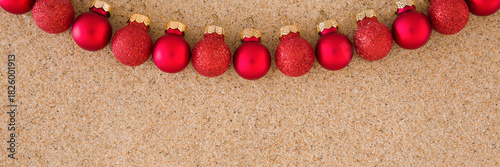 Beautiful shiny red baubles on light brown dry sea sand background. Closeup. Christmas decoration. Wide banner. Empty place for text. Top down view.