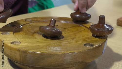 Small wooden traditional Malay spinning tops are spun on a wooden platform that slopes to the center. Multiple tops are competed in this activity, representing a vanishing traditional Malay game.
