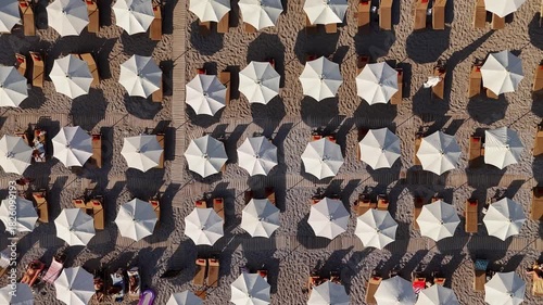 Aerial top view of neatly arranged beach umbrellas and sunbeds on a sandy shore. Symmetrical summer setup with people relaxing under bright sunlight. Perfect vacation and resort atmosphere.
