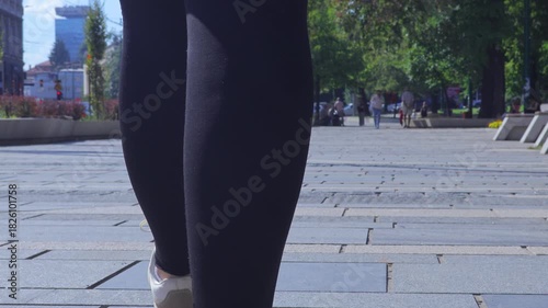 Low-angle rear view of a person wearing sneakers and black pants walking across a paved urban square on a sunny day, with pedestrians and trees in the background.