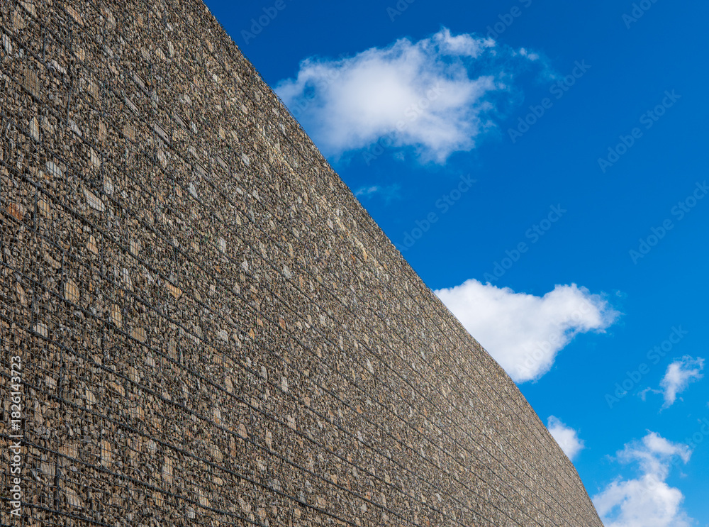 custom made wallpaper toronto digitalLarge gabion retaining wall filled with stones, built for slope protection and erosion control, photographed against a blue sky.