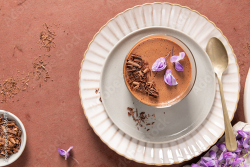 An overhead view features one creamy chocolate mousse in a glass, styled on plates with a spoon, a bowl of shavings, and purple flowers on a rustic surface.