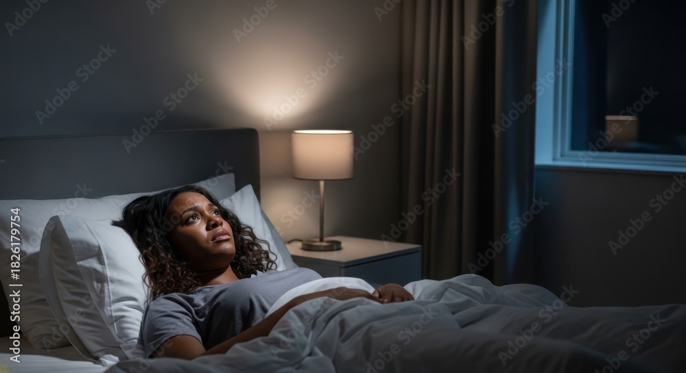 Fototapeta premium Woman lying awake in bed at night suffering from insomnia. Sleepless night and mental health. Bedroom with lamp and window, dark setting.