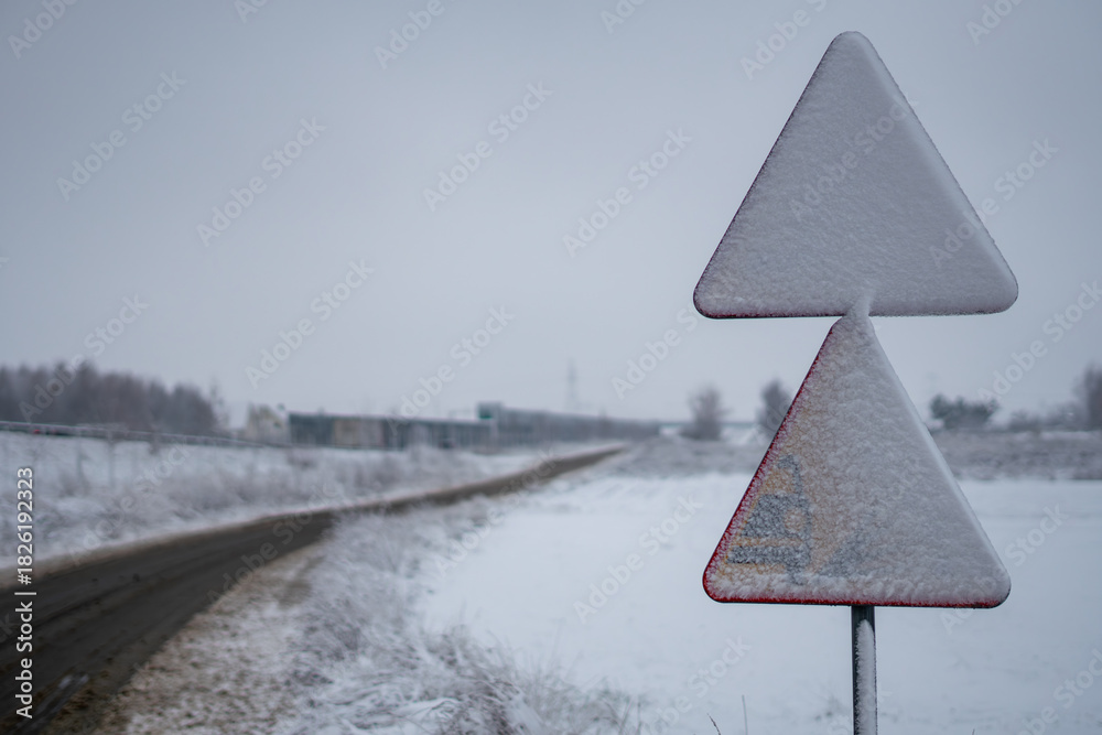 Fototapeta premium Snow covered warning road sign beside rural road in winter landscape with open fields background