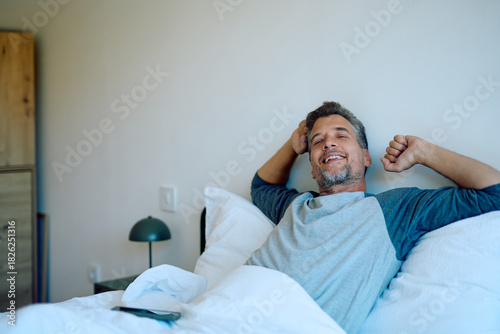 Tablou pe pânză Mature man happily stretching and smiling in bed after a good night's sleep, fee