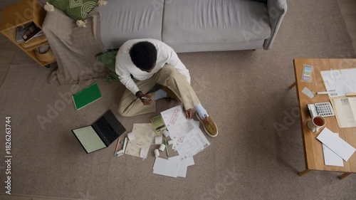 Top down view shot of African American man organizing receipts and unpaid bills on floor, calculating monthly expenses with calculator at home, showing financial concern
