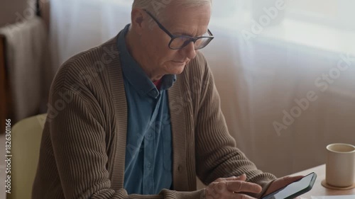 Tilt up of elderly Caucasian man checking bills and managing finances using smartphone at wooden desk in home setting