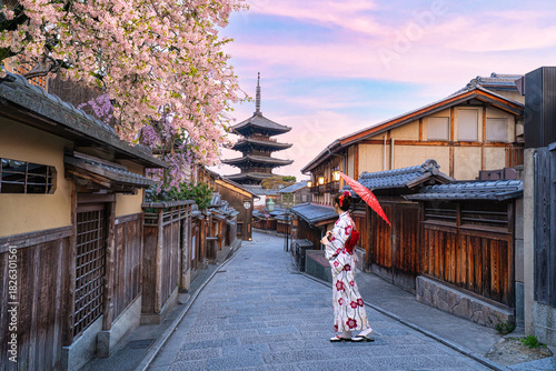 A young Japanese woman in a traditional kimono stands beside the Yasaka Pagoda in Kyoto, Japan, amidst blooming cherry blossoms.