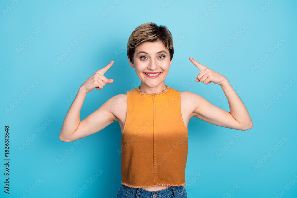 Fototapeta premium Happy stylish young woman with short blonde hair pointing to her head against blue background in casual chic outfit