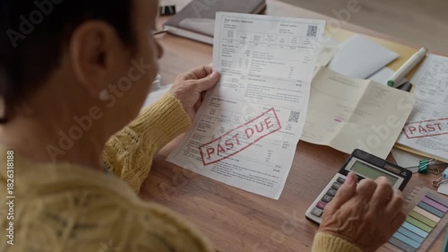 Over the shoulder shot of elderly woman sitting at home desk holding overdue bill while using calculator to manage household finances