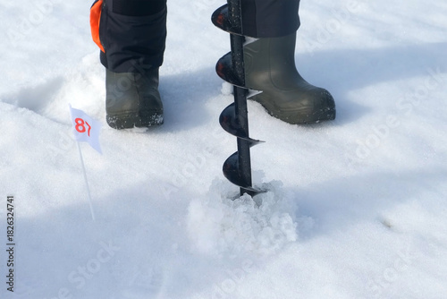 Ice fisherman drilling hole in lake ice with ice auger preparing to catch fish in winter. Man fishing on frozen river with ice and snow. Outdoor winter activity hobby leisure recreation kind of sport.