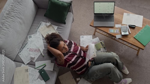 Top down view shot of worried Biracial woman sitting on floor at home surrounded by stacks of overdue bills using laptop to manage finances and plan payments