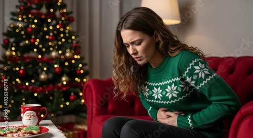 Woman with stomach pain in an ugly Christmas sweater sitting on a couch near a festive tree. Concept of holiday discomfort or food poisoning.