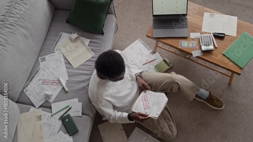 Top down view shot of depressed Black man sitting on floor among stacks of overdue bills, leaning his head back on sofa, showing stress, worry, and emotional burden of debt