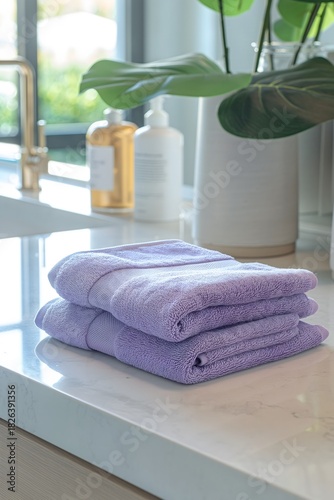 Two plush purple towels are stacked neatly on a smooth kitchen countertop. A bright and airy space features green plants and clear bottles in the background, creating a fresh atmosphere