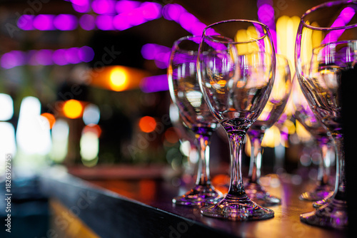 Empty clear wineglasses stand in rows on bar counter at nightclub, blurred purple neon and yellow lights on background. Clean cocktail glasses ready for celebrate party.