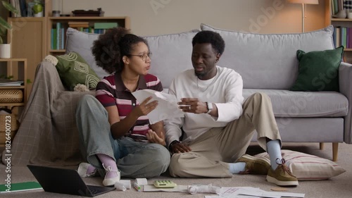 Emotional Biracial couple sitting on living room floor arguing while analyzing bills and household expenses, showing financial stress and tension in realistic home setting
