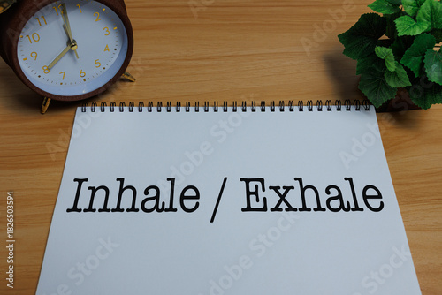 A spiral notebook with the text “Inhale / Exhale” placed on a desk beside an alarm clock, symbolizing a simple wellness practice of mindful breathing and stress relief.