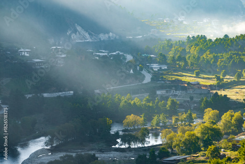Aerial view of sunbeams filtering through the morning mist, casting a golden glow over the winding Chhudzom-Haa Road and lush valley, Haa Valley, Paro, Bhutan.