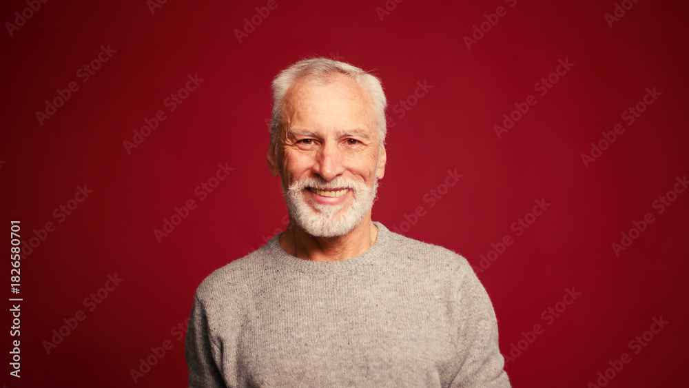 Obraz premium Portrait of happy senior man touching grey beard looking at camera posing isolated on red background