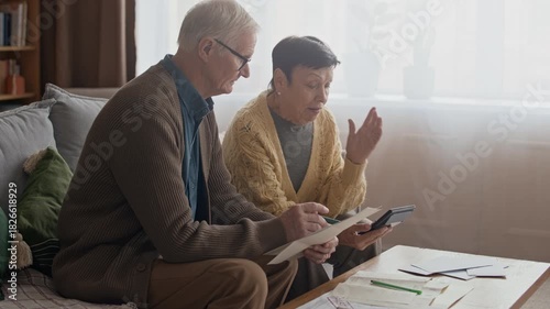 Medium shot of elderly Caucasian couple calculating household expenses and bills on sofa using calculator, dealing with financial difficulties in cozy living room at home