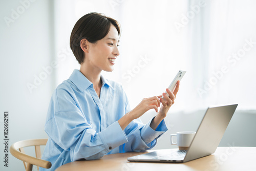 リビングで椅子に座り、パソコンを開いて携帯電話を見る日本人女性