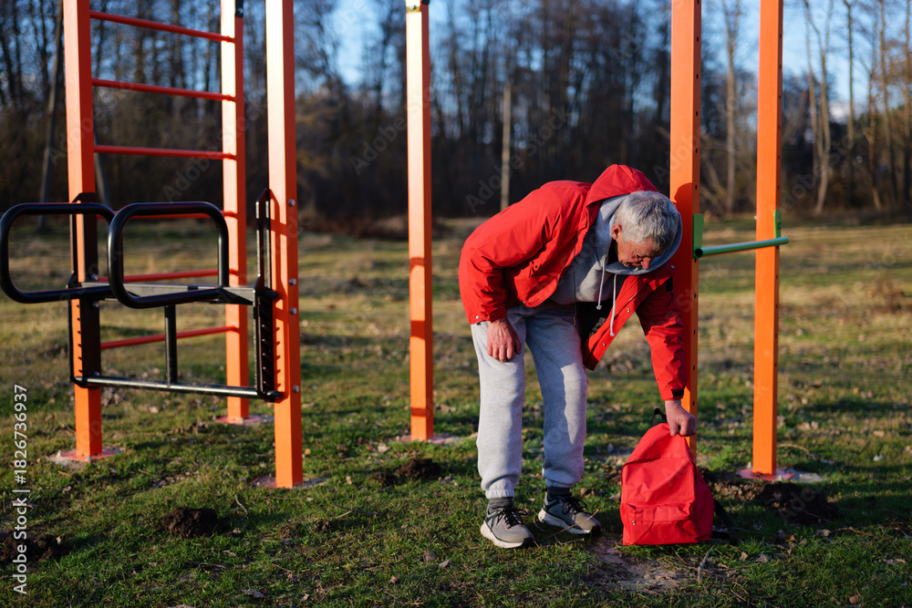 Fototapeta premium An older man wearing a red jacket bends down to pick up a red bag near outdoor gym equipment in a park, surrounded by trees on a sunny afternoon