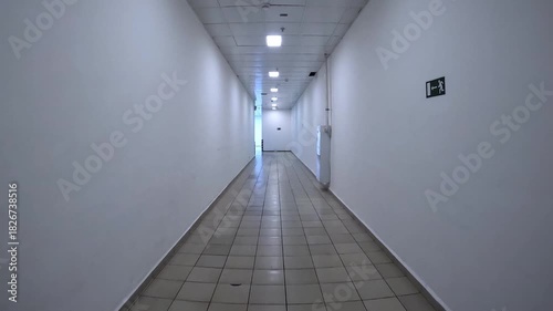 Long, narrow corridor with textured white walls, tiled floor, and fluorescent lighting. Backstage. This creates sense of anxiety and disorientation, confinement, isolation. Slow, forward movement.