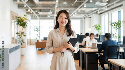 Portrait of a smiling Asian businesswoman in a beige dress holding a tablet, standing in a modern open-plan office with colleagues working in the background