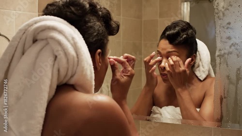 Woman applying skincare product to face reflection in bathroom mirror after shower with towel wrapped head