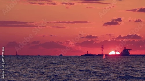 Number 8. Dramatic red-orange sunset, with the outline of coastline and ships and sailboats floating on water visible on horizon.