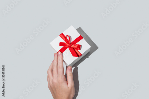 A hand holding a white gift box with a red ribbon on an isolated light background