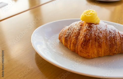 Primer plano macro de croissant o medialuna horneada, cubierta con copo de crema pastelera y azúcar impalpable, servida en plato blanco sobre mesa de madera.