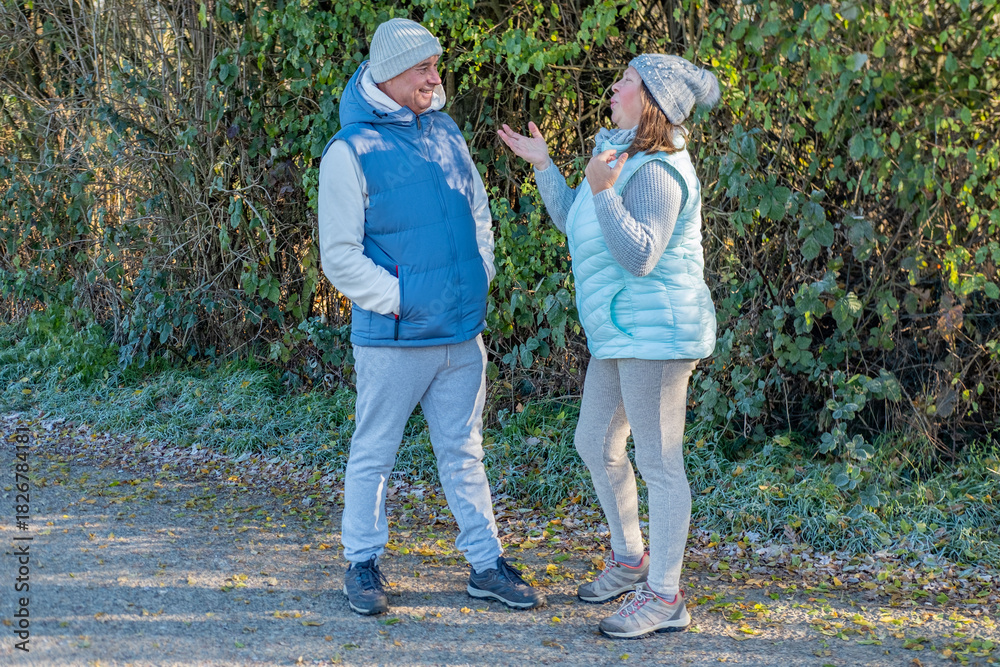 Fototapeta premium two adults in warm clothes talking outdoors near green bushes, man smiling while woman gestures energetically during walk, outdoor communication, active conversation, friendly communication moment