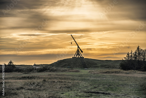 Fototapeta Naklejka Na Ścianę i Meble -  Silhouette of a Historic Trebuchet on a Hill at Sunset Over Open Grassland Landscape, Skagen, Denmark