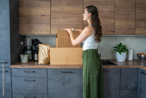 Young woman unpacking a box with pre-portioned ingredients for cooking on the kitchen counter. Concept of meal kit delivery service and easy home cooking with fresh food.