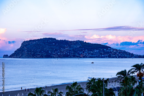 Fototapeta Naklejka Na Ścianę i Meble -  Alanya’s historic peninsula with ancient fortress on red cliffs, evening Beach promenade with palms, tropical beach, historic peninsula Alanya, seaside view palm trees, resort locations, ocean waves