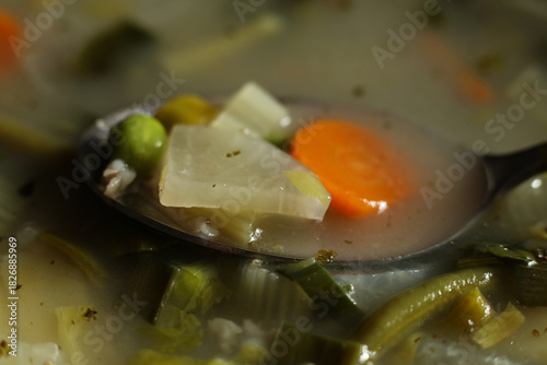 Macro close-up of a spoon filled with vegetables in clear broth. Visible peas, carrots, celery, and grains captured with shallow depth of field and warm natural light. Detailed food texture