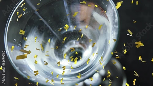 Hand entering upper right stirring glass, forming vortex bubbles, olive dropping, confetti drifting