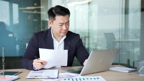 Confused puzzled asian businessman having difficulty with paperwork sitting in business office. Frustrated financier unhappy with bad financial results reviewing documents and looking at laptop screen