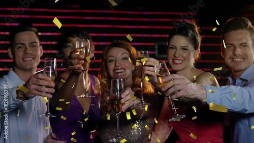 Group of five raising flutes, releasing gold confetti, sipping, celebrating event by LED backdrop