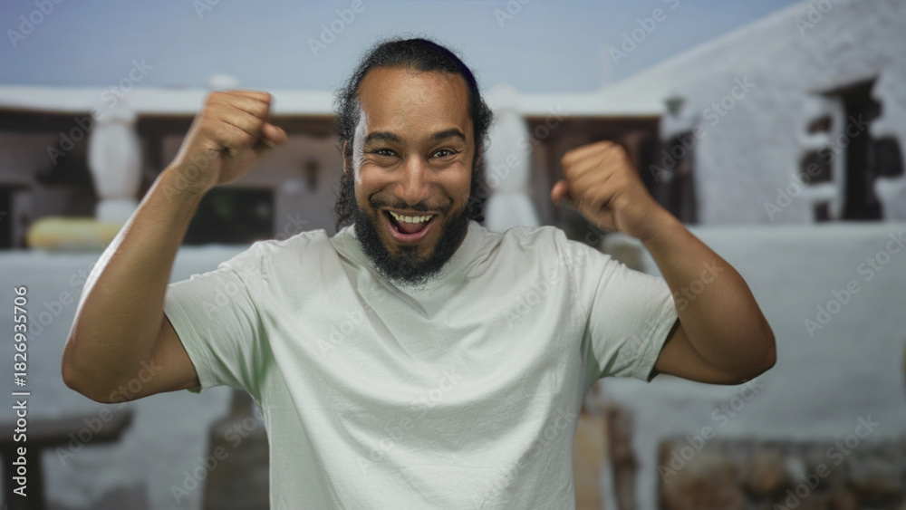 Naklejka premium Man raises fists in celebration at a building entrance, smiling widely with bare forearms and white tshirt, ponytail and beard visible; victory joy.