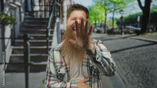 Картината върху платно Man with long beard and plaid shirt pointing finger toward camera in urban street with building facade visible; amusement