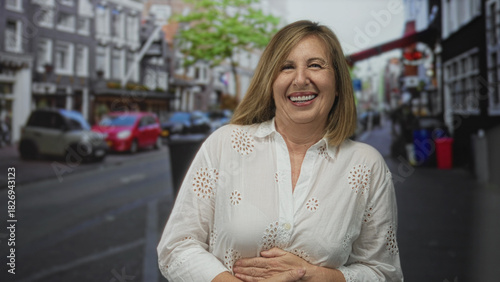 Woman smiling broadly while clasping her stomach in a bustling city street with blurred cars and storefronts; happiness.